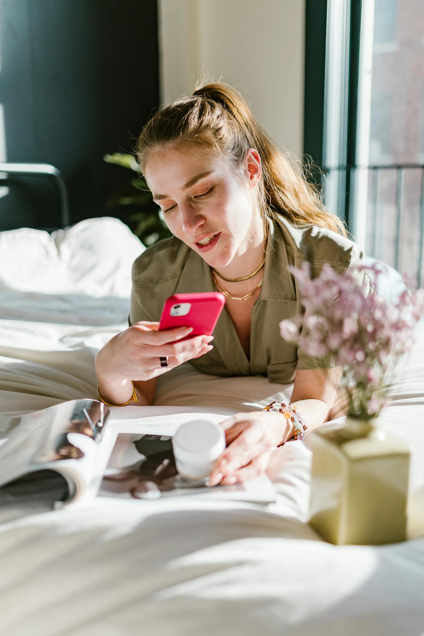 Woman lying in bed using her smartphone with flowers in the foreground and magazine nearby.